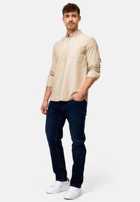 Beige Hemd mit Kragen, langen Ärmeln und Brusttasche, kombiniert mit dunkelblauen Jeans und weißen Sneakern. Glatte Textur.