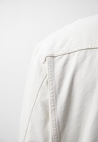 Nærbillede af en hvid denimjakke skulder, der viser detaljeret syning og stofstruktur på en enkel hvid baggrund.