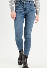 Højfløjede, tætsiddende blå jeans i strækdenim med subtilt slid, med fem lommer og lynlås. Kombinationen med sorte ankelstøvler.