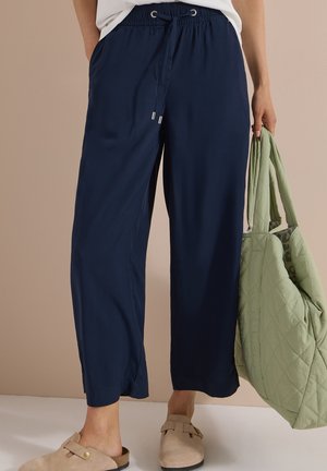 Person iført marineblå snorbukser og beige slip-on sko, der holder en stor lysegrøn quiltet taske i den ene hånd.