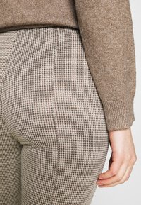 Brauner Strickpullover über enganliegenden, hounds-tooth-musterierten Hosen in Beige und Braun. Die Hose zeichnet sich durch einen strukturierten Stoff und eine maßgeschneiderte Passform aus.