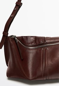 Sac bandoulière en cuir marron avec fermeture éclair argentée et sangle réglable ornée de clous en métal sur fond blanc.