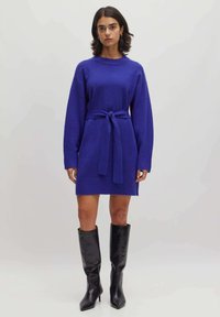 Gebreide jurk in een levendige blauwe kleur met lange mouwen, een ceintuur in de taille en een rechte pasvorm. Gecombineerd met zwarte leren laarzen tot de knie.