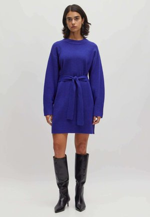 Strickkleid aus leuchtendem Blau mit langen Ärmeln, tailliertem Schnitt und geradem Fall. Kombiniert mit kniehohen schwarzen Lederstiefeln.
