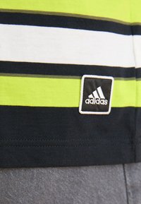 Zaļa un melna svītraina auduma t-krekls ar baltu svītru, ar izšūtu melnu un baltu Adidas logo uz apakšmalas.