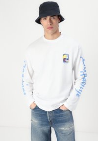 Tricou alb cu mânecă lungă, având un imprimeu grafic multicolor pe piept și text albastru pe mâneci; asociat cu blugi albaștri distruși.