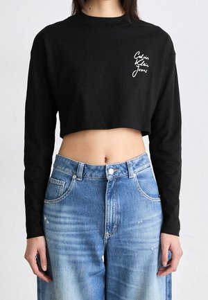 Kobieta w czarnym skróconym długim topie z białym logo "Calvin Klein Jeans" i jasnoniebieskich jeansach z wysokim stanem.