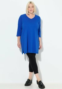 Blaue Tunika mit V-Ausschnitt, dreiviertel Ärmeln und gefälteltem Vorderteil, kombiniert mit schwarzen Leggings und klobigen schwarzen Schuhen. Glatte Textur.