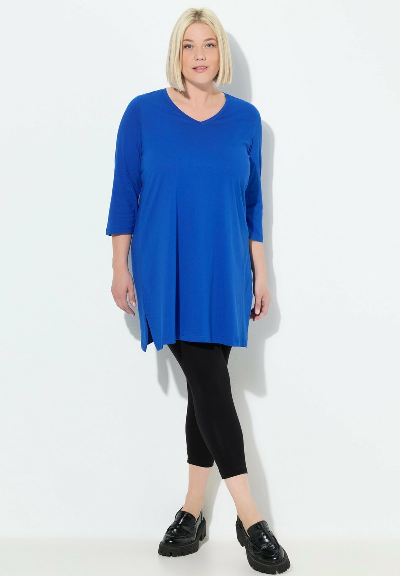 Blaue Tunika mit V-Ausschnitt, dreiviertel Ärmeln und gefälteltem Vorderteil, kombiniert mit schwarzen Leggings und klobigen schwarzen Schuhen. Glatte Textur.