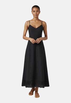 Mujer de pie con un vestido midi negro sin mangas con tirantes finos y dobladillo festoneado, combinado con sandalias de tiras marrones.
