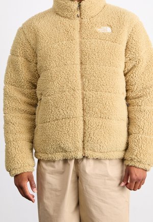 Beige flauschige Fleecejacke mit Frontreißverschluss und zwei Reißverschlusstaschen, getragen mit hellbeiger Hose, mit The North Face Logo auf der Brust.
