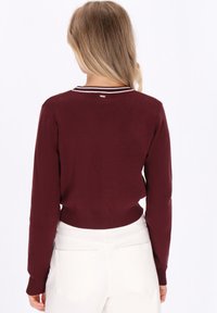 Blonde woman wearing a burgundy long-sleeve sweater with white striped collar, paired with high-waisted white pants, facing away.