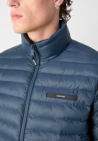 Giubbotto imbottito navy con colletto alto, trapuntatura orizzontale e zip frontale. Presenta un'etichetta nera con la scritta "Calvin Klein" sul petto.