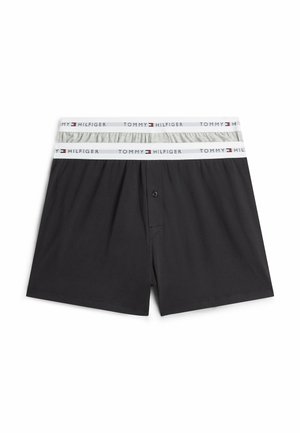 Junger Mann, barfuß stehend vor weißem Hintergrund, trägt schwarze Tommy Hilfiger Boxershorts mit Logo am Bund.