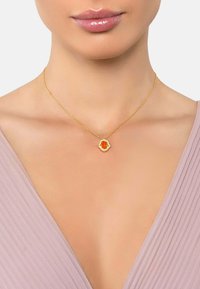 Collana d'oro con un pendente floreale dal centro arancione, appesa a una delicata catenina, indossata sopra un top di un leggero colore viola.