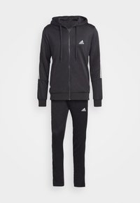 Veste zippée noire avec capuche et logo blanc, associée à un pantalon noir assorti. Présente des bandes blanches sur les manches et une texture lisse.