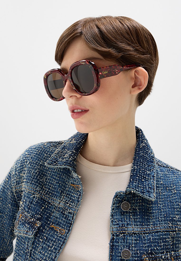 Sonnenbrille mit großen, runden Rahmen im Schildpattmuster, dunklen Gläsern und Branding an der Seite; getragen mit einer strukturierten Jeansjacke.