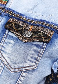 Ljusblå jeansjacka med en svart spetsfickdetalj med paljetter och en dekorativ knapp. Fransiga kanter och sömnadsdetaljer syns.