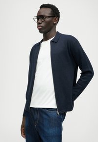 Casaco de malha azul-marinho com fecho, com gola, combinado com uma t-shirt branca e jeans de ganga escura. Textura subtil e design minimalista.