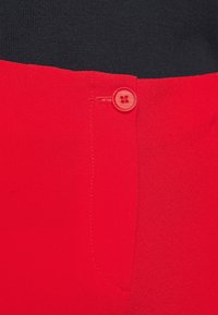 Pantalon rouge avec une texture lisse, un unique bouton rouge à la taille et une poche avant avec des coutures nettes. L'arrière-plan est en tissu noir.