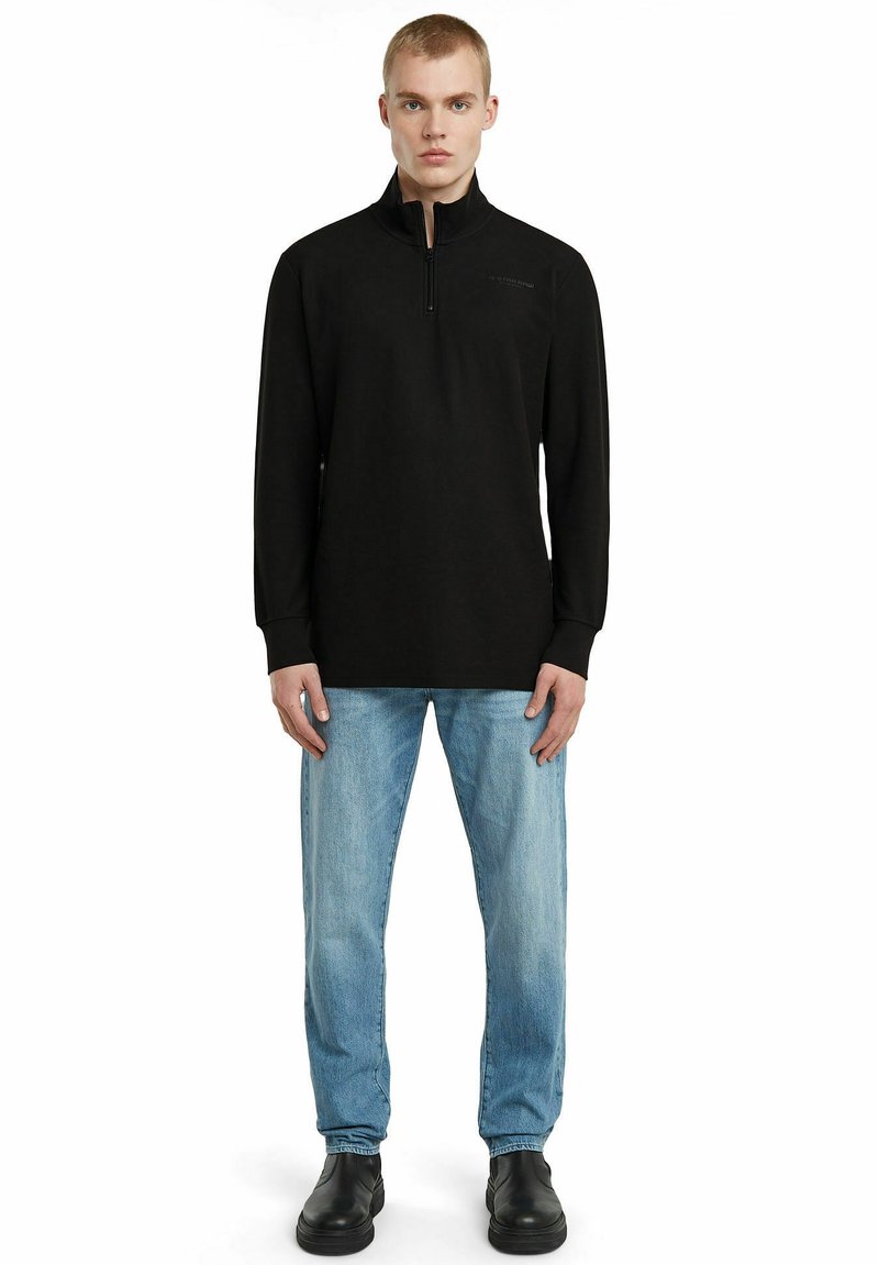 Schwarzer langärmeliger Pullover mit halbem Reißverschluss aus weichem Material, mit hohem Kragen und dezentem Branding, kombiniert mit hellblauen Jeans und schwarzen Schuhen.