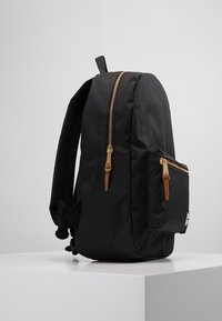 Zwarte rugzak met een strak ontwerp, voorzien van een gouden rits en beige leren accenten. Heeft één hoofdvak en een voorvak.