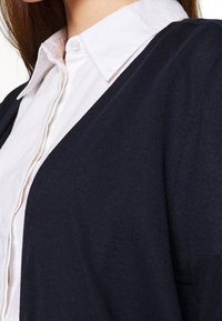 Navy cardigan over a white button-up shirt, featuring a textured fabric with a V-neck design, visible collar, and soft drape.