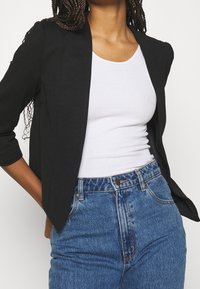 Persoon gekleed in een zwart blazer over een witte mouwloze top, ingestopt in hoog-wijde blauwe denimjeans, staand met één hand op de heup.