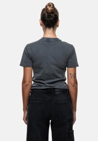 T-shirt in cotone grigio con maniche corte, caratterizzato da un collo rotondo e un sottile dettaglio del logo sul retro. Indossato con jeans neri a vita alta.