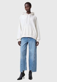 Sudadera blanca con un bolsillo frontal y cordones, combinada con jeans de pierna ancha azul claro. Botas negras hasta el tobillo completan el atuendo.