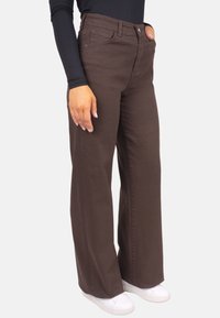 Pantalon large marron en mélange de coton, avec poches avant et braguette zippée, associé à des baskets blanches et un haut noir ajusté.