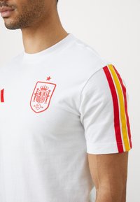 Λευκό βαμβακερό t-shirt με κόκκινο έμβλημα ισπανικής ποδοσφαιρικής ομάδας και δύο κόκκινες και κίτρινες ρίγες στα μανίκια. Κλασικός σχεδιασμός με στρογγυλή λαιμόκοψη.