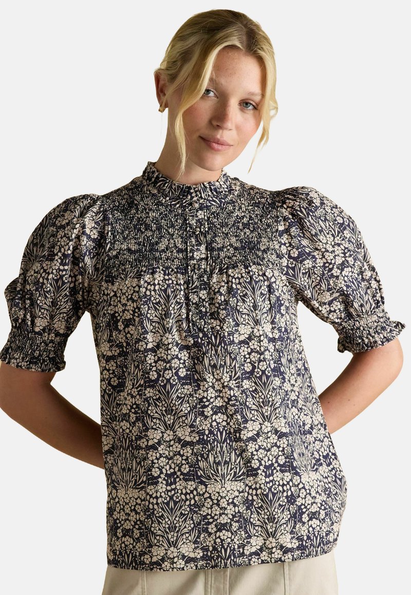 Ziedu apdrukas blūze ar augstu kakla izgriezumu, pūkainām piedurknēm un smalku virsotni, kas ir tumši zila ar krēmkrāsas akcentiem; viegla auduma.