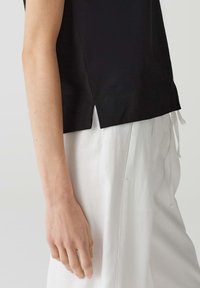 Zwarte top met korte mouwen en een ronde hals met zijsplitten, gedragen over witte, losse broek. De stof lijkt glad.