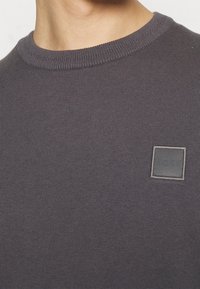 Grå stickad tröja med rund hals och ribbade ärmslut. Har en rektangulär BOSS-logotyp på bröstet. Slät textur.