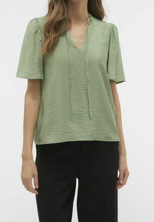 Vrouw draagt een lichtgroene gestructureerde korte mouwen blouse met een V-hals en hangende strikjes, gecombineerd met een zwarte broek, staand tegen een witte achtergrond.