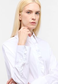 Blusa bianca con dettagli a volant sottili lungo la placket e i polsini, caratterizzata da un colletto classico e polsini abbottonati in un tessuto morbido.