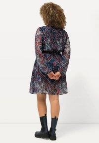 Häikäisevä, kuviollinen mekko tummansinisessä, punaisessa ja harmaassa värissä, pitkillä hihoilla ja smockattulla selällä. Yhdistetty polven yli ulottuviin mustiin kumisaappaisiin.
