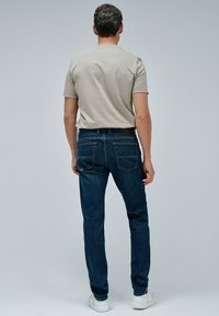 Hombre vistiendo una camiseta de manga corta beige, jeans oscuros y zapatillas blancas, de pie, de espaldas, frente a un fondo gris liso.