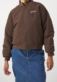 Osoba oblečená v hnědém zipovém svetru značky New Balance, pod kterým má tigrovaný rolák a modrou džínovou sukni, s viditelnou rukou a zápěstím.