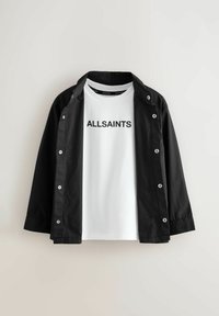 Melna pogca jaka, kas kārtota virs balta T-kreklā ar "ALLSAINTS" tekstu centrā uz krūškurvja, attēlota uz gaiša fona.