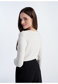 Biały dzianinowy sweter z długimi rękawami, okrągłym dekoltem i złotymi guzikami na ramionach, zestawiony z czarnymi spodniami o wysokim stanie.