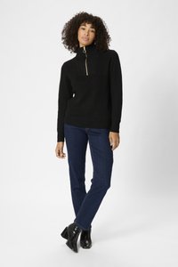 Pull en tricot noir avec demi-fermeture éclair et col haut, associé à un jean en denim bleu et des bottines à talons épais noires.