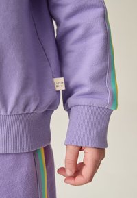 Lila Sweatshirt mit gerippten Bündchen und Saum, mit einem Etikett, auf dem "Little Bird" steht. Seitliche Streifen in Grün, Gelb und Rot. Glatte Textur.