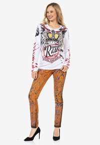 Chemise blanche à manches longues avec un design de couronne graphique et des accents rouges, associée à un jean skinny orange avec des éclaboussures bleues et des détails de texte.