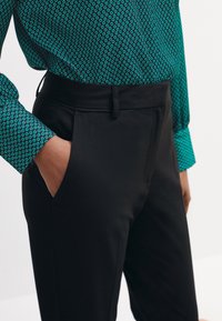 Czarne dopasowane spodnie z ukośnymi przednimi kieszeniami, noszone z zieloną bluzką w wzory z geometrycznym motywem. Gładki, dopasowany materiał.