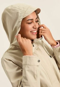 Jachet�ă impermeabilă bej cu glugă, dotată cu un fermoar frontal, șnur elastic și detalii de cusătură vizibile. Căptușeală interioară moale.