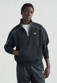 Μαύρο φούτερ με φερμουάρ και λευκή επένδυση, με μπροστινή τσέπη, που φέρει μικρό λογότυπο Nike στο στήθος. Συνδυάζεται με μαύρο παντελόνι.