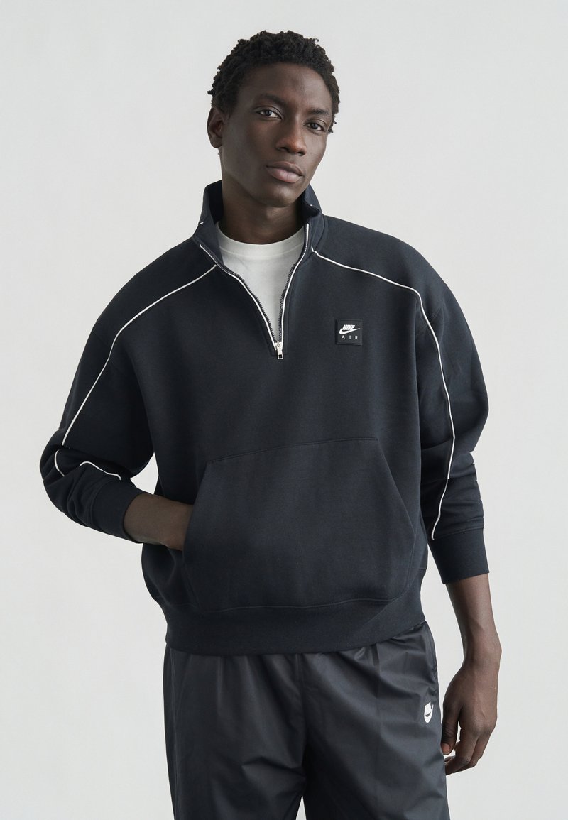 Μαύρο φούτερ με φερμουάρ και λευκή επένδυση, με μπροστινή τσέπη, που φέρει μικρό λογότυπο Nike στο στήθος. Συνδυάζεται με μαύρο παντελόνι.