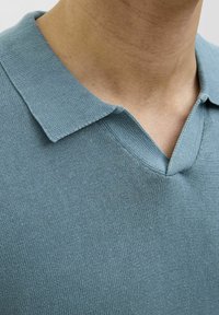 Polo shirt bleu en tricot avec un design en col en V, col court et tissu texturé. Un gros plan met en évidence les détails des coutures.